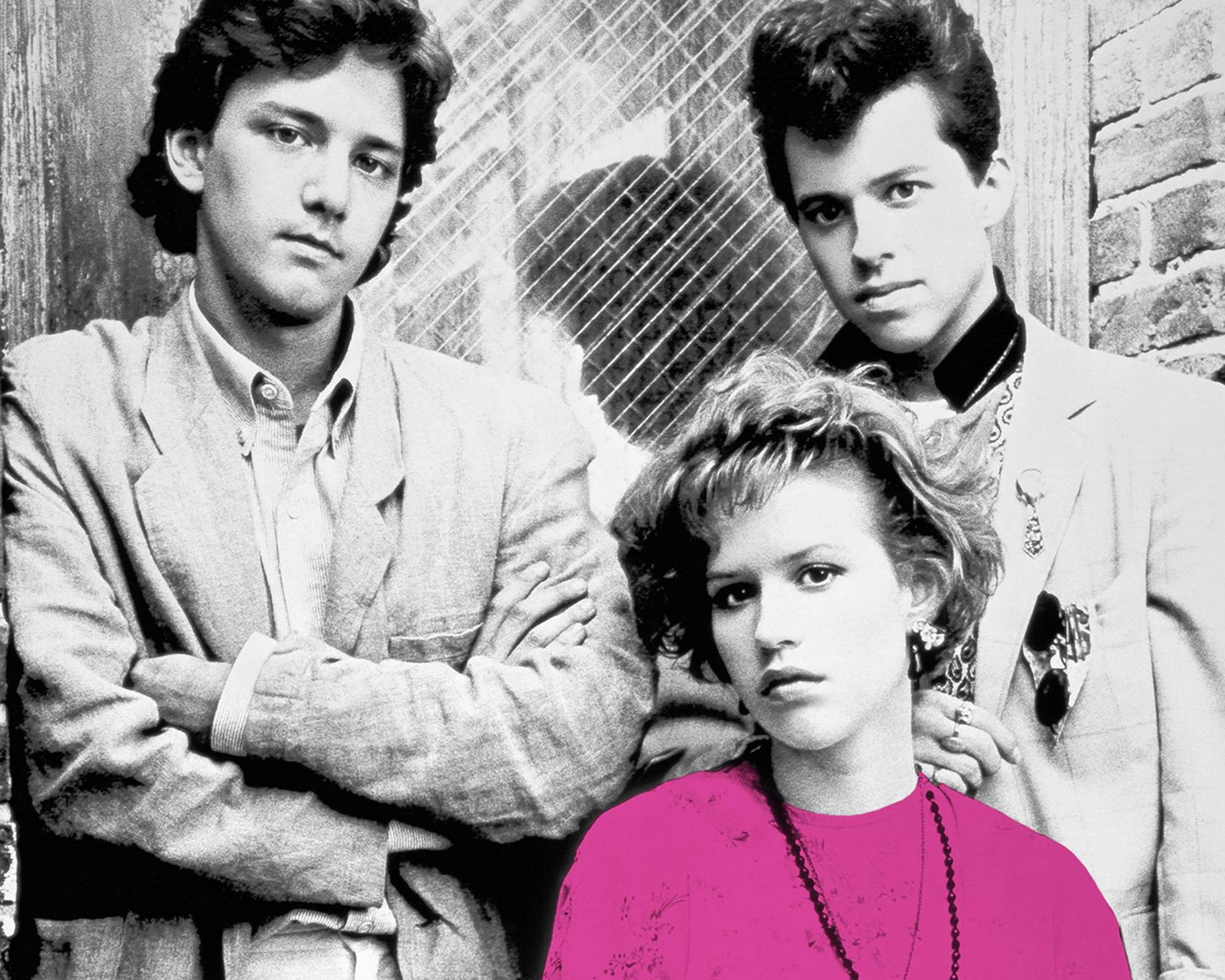 Black and white portrait of three young adults posed together against a rustic wall with a chain-link fence, with the woman in the foreground wearing a bright pink top.