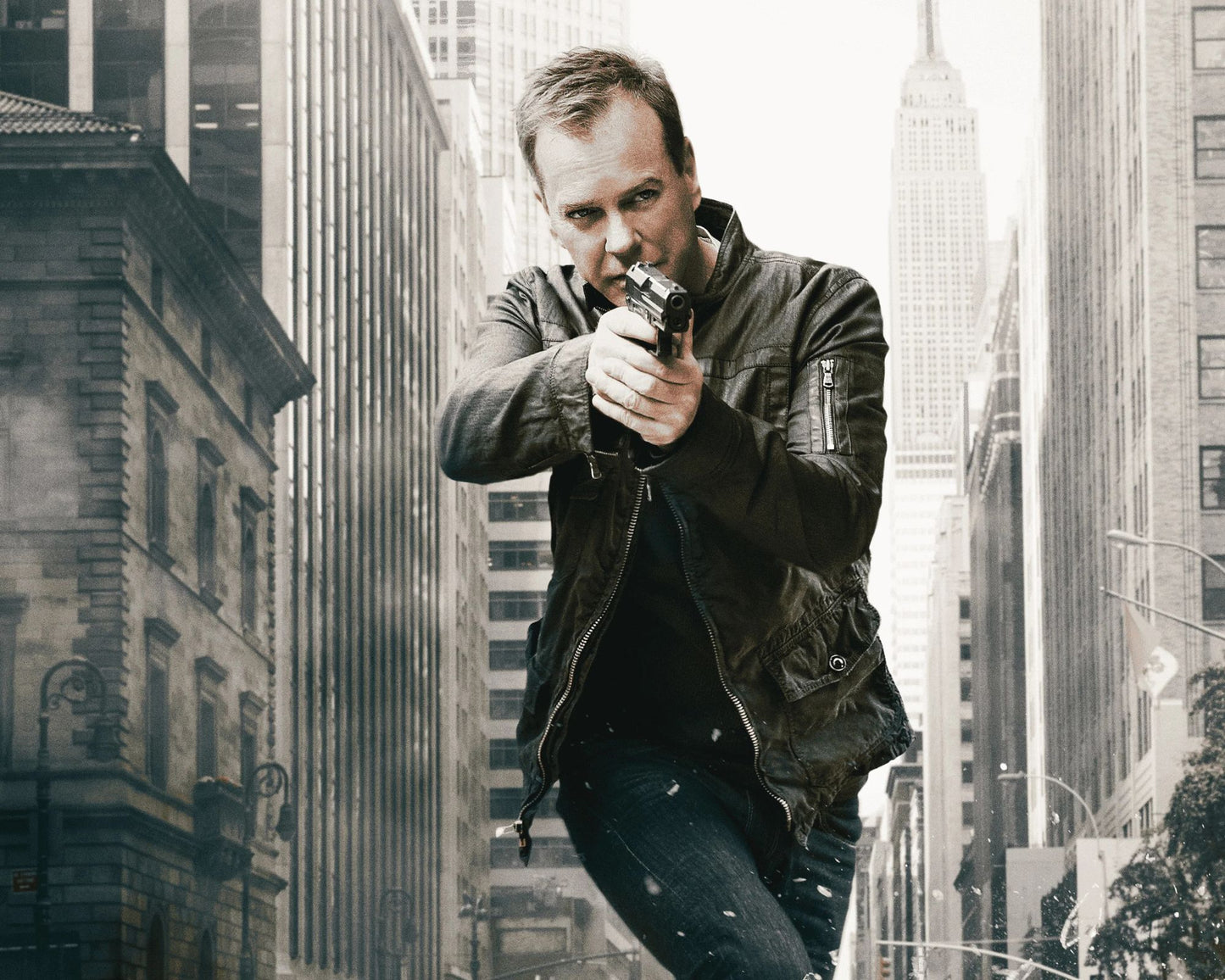 A man in a dark leather jacket and jeans aims a handgun down a city street lined with tall buildings.