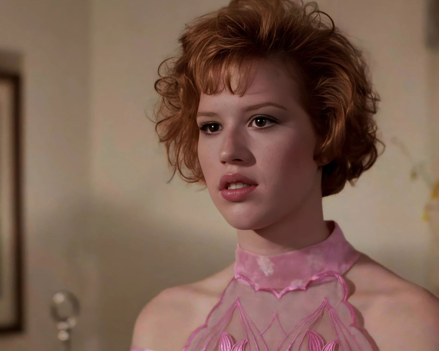A close-up portrait of a woman with short curly auburn hair wearing a pink sheer lace dress, set against a softly lit beige indoor background.