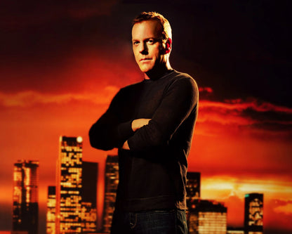 A man in a black long-sleeve shirt and jeans stands with arms crossed against a dramatic orange and red sunset city skyline background.
