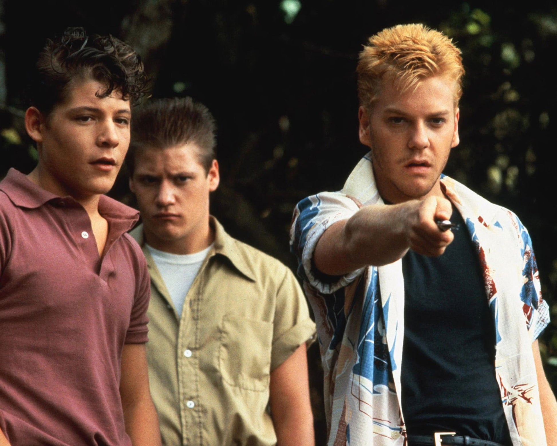 Three young men standing outdoors in front of dark foliage, with the man on the right pointing forward while wearing a white shirt with blue and red graphics over a dark undershirt.