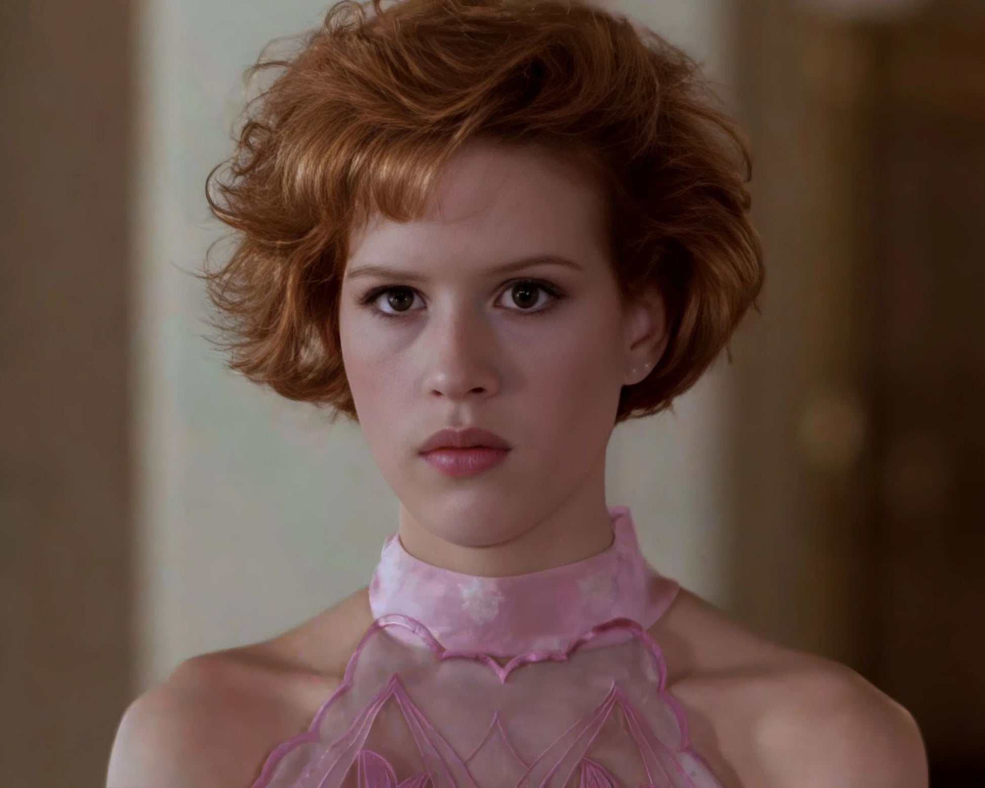 A close-up portrait of a woman with short, wavy red hair wearing a sheer pink high-neck top with a floral pattern.