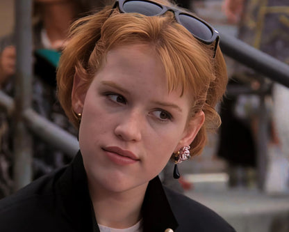 A close-up shot of a woman with short auburn hair, sunglasses on her head, and a black jacket, wearing a pink and black earring. The background is a blurred outdoor scene with people and metal railings.