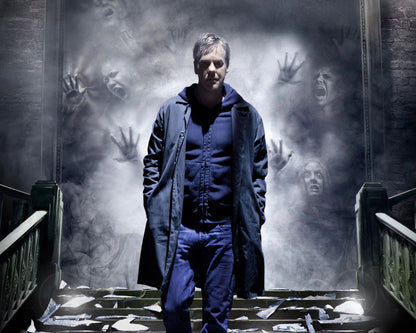 A man in a dark navy blue trench coat and matching blue shirt and jeans stands on a staircase in a dim, smoky hall, with ghostly faces and hands projected onto the wall behind him.