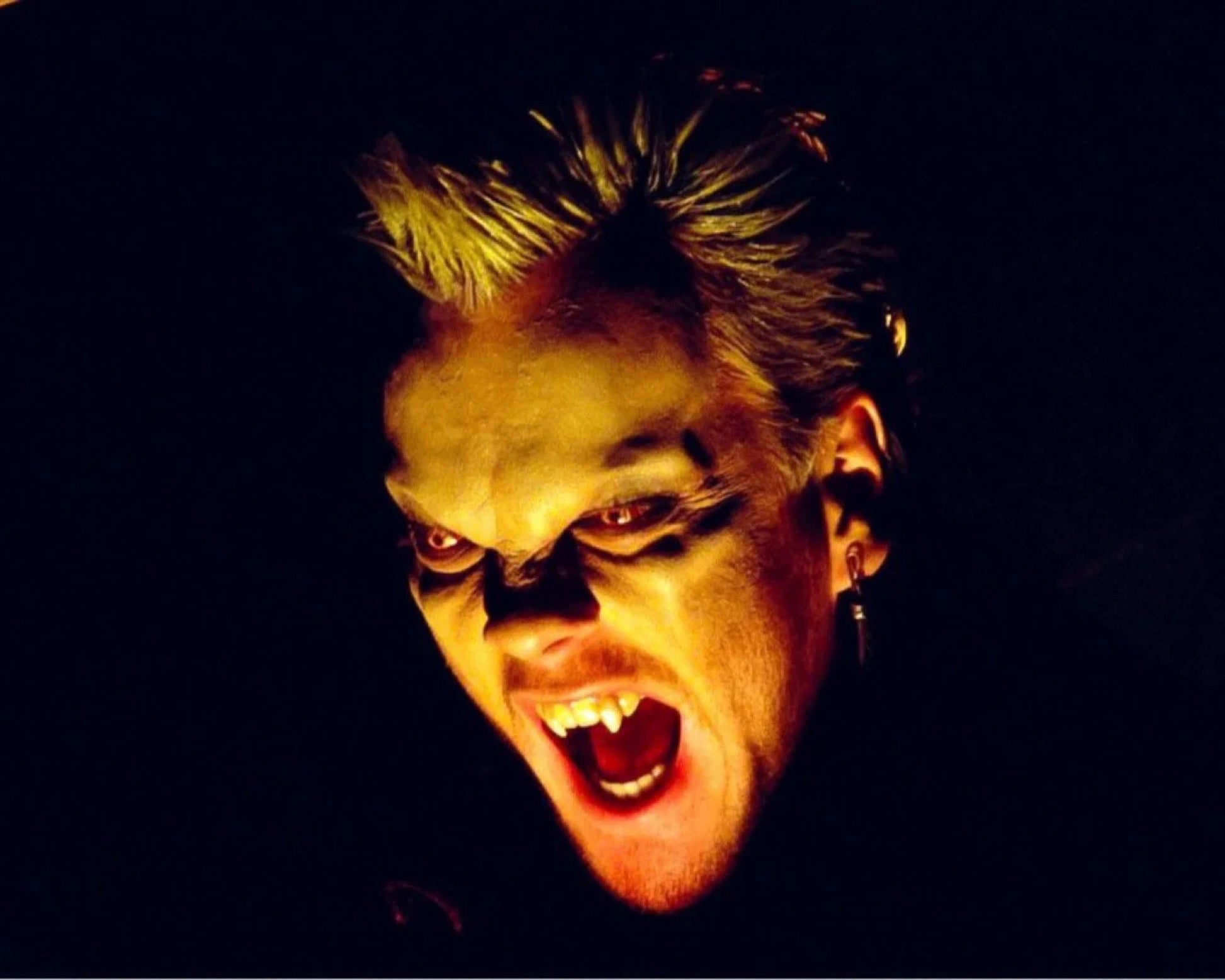 A close-up portrait of a person with dramatic vampire-style makeup, including pale skin, dark eye makeup, and fangs, lit by a warm yellow light against a black background.