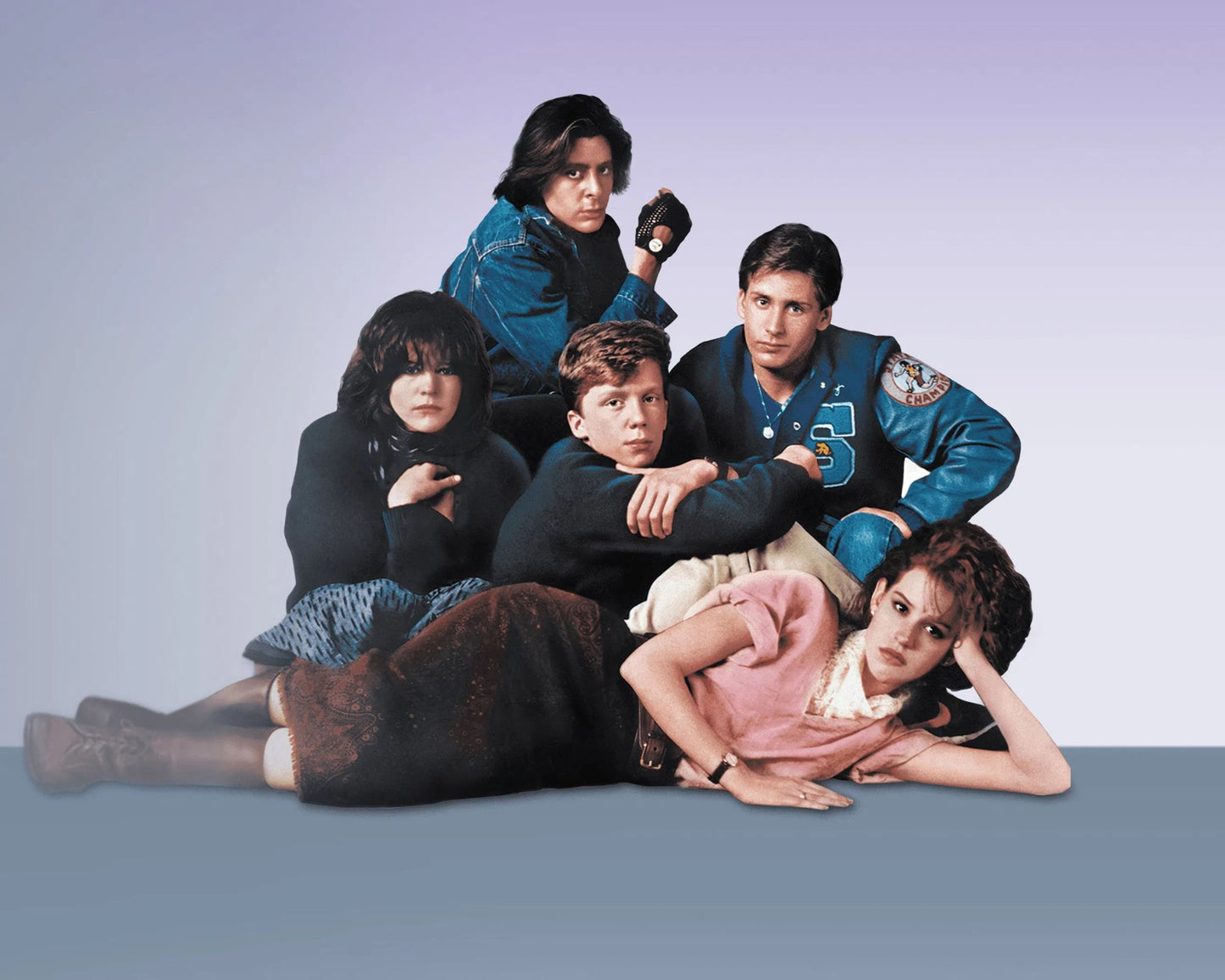 A group of five people posed together against a soft lavender-to-blue studio background. The scene appears to be a movie or TV still, with one person in a blue jacket at the top, two people in dark clothing in the center, and two people reclining at the bottom.
