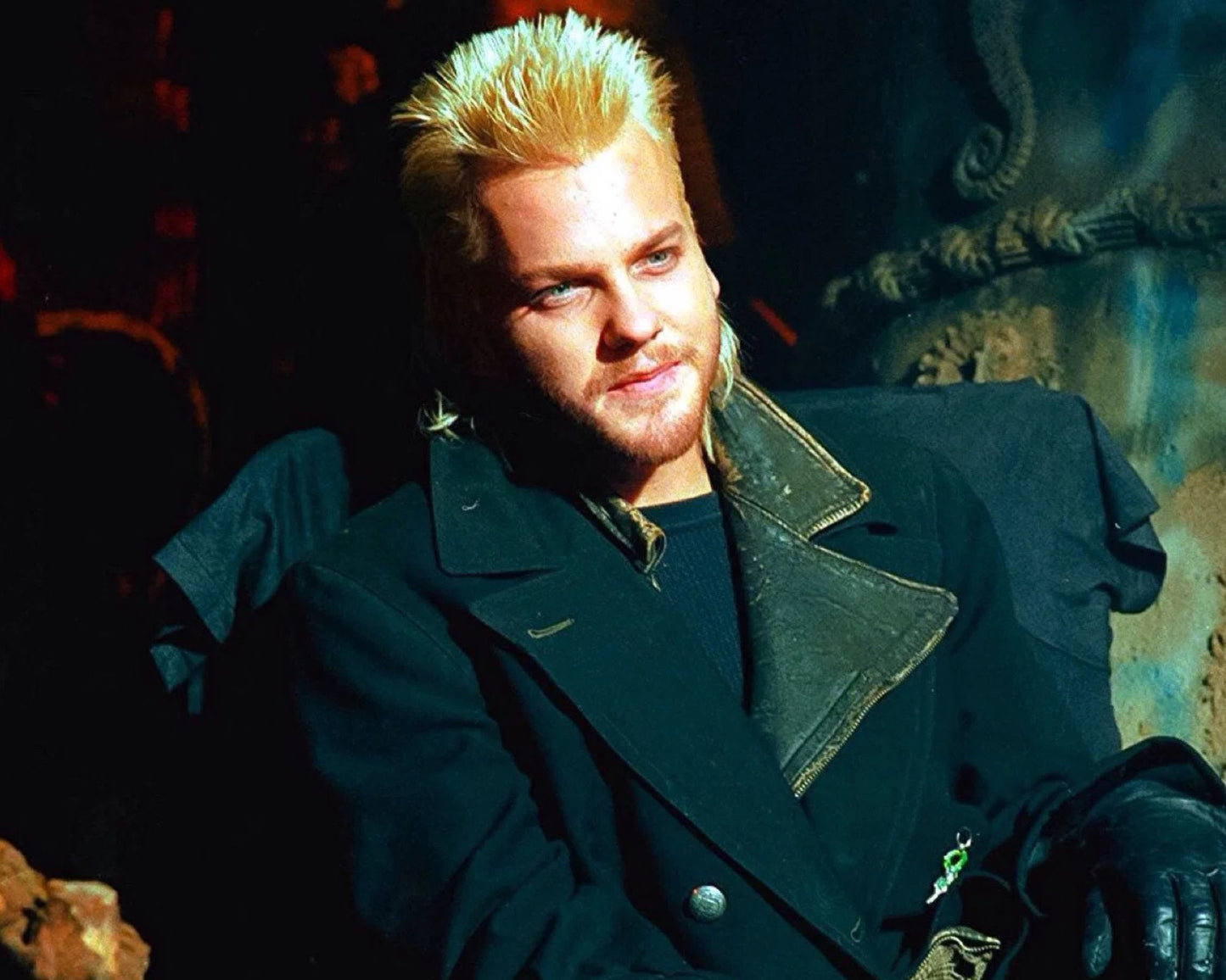 A man with spiky blond hair wearing a dark coat and black gloves, seated in a dimly lit setting with dramatic lighting.