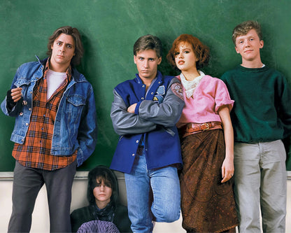 Four young adults and a woman posing together in front of a green chalkboard, dressed in casual 1980s style clothing including denim jackets, a varsity jacket, a pink blouse, and a brown skirt.