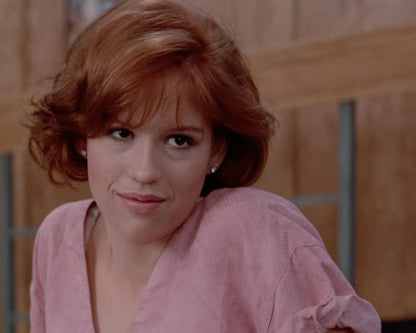 A close-up shot of a woman with short red hair wearing a light pink top, looking directly at the camera against a warm-toned wooden background.
