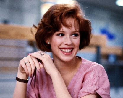 A smiling woman with short auburn hair wearing a pink top, photographed indoors with a softly blurred background.