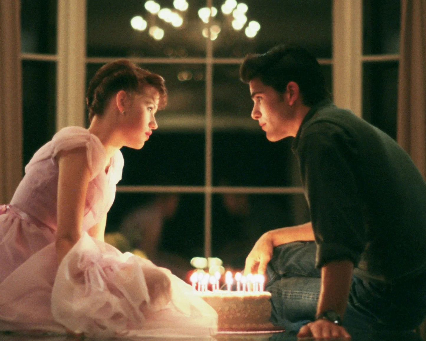 A man and a woman in a dimly lit room, kneeling down to look at a birthday cake with lit candles. The woman is wearing a light pink dress, and the man is wearing a dark green shirt and jeans.