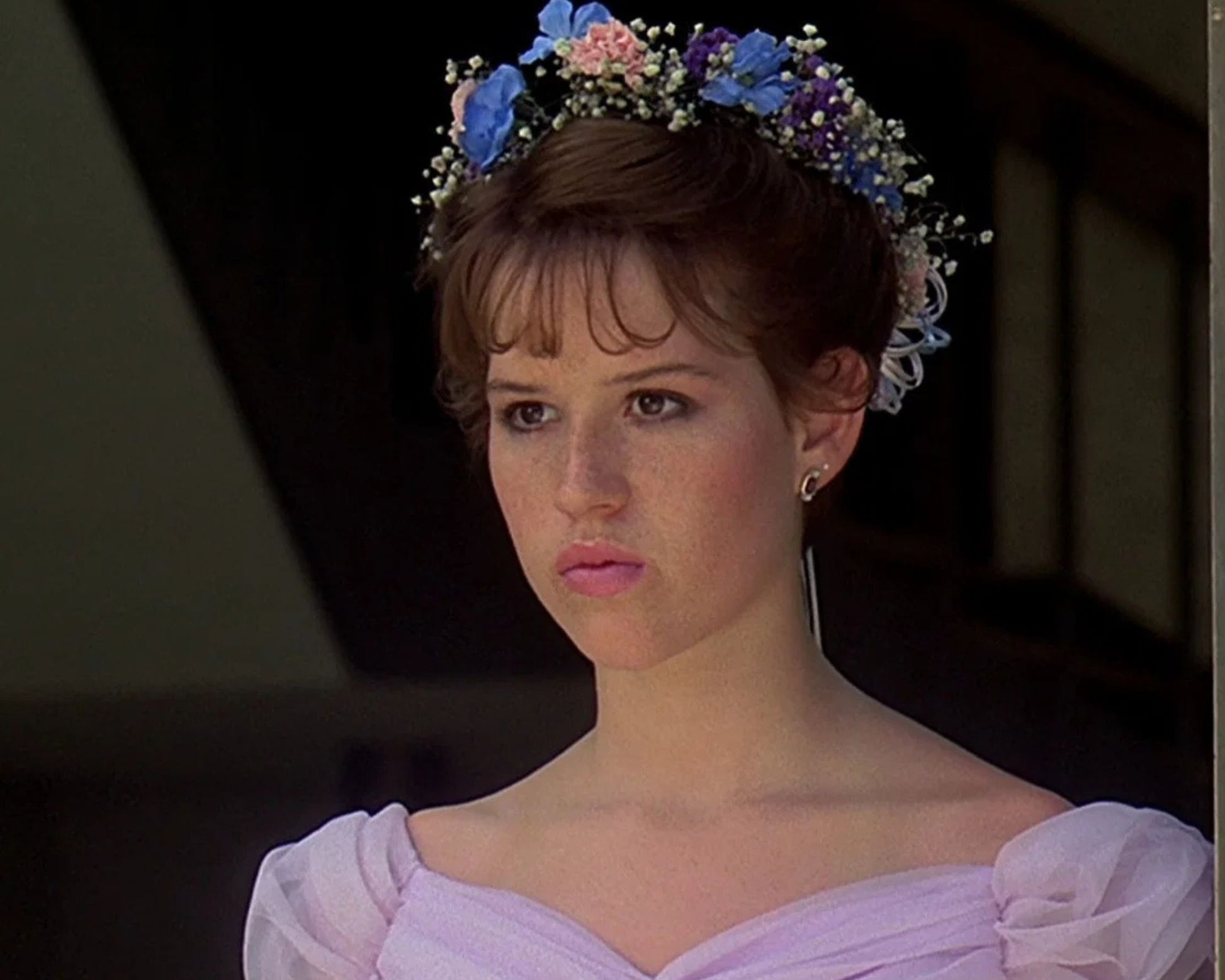 A close-up portrait of a woman with short brown hair wearing a floral crown of blue, pink, and white flowers, dressed in a pale lavender off-the-shoulder gown. The background is a dark, shadowed interior.