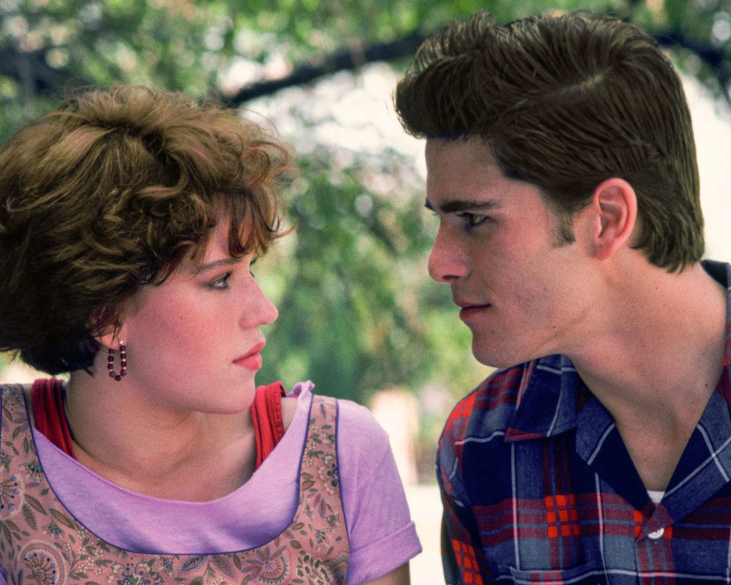 A close-up shot of a young couple looking at each other outdoors. The woman on the left has short curly brown hair and wears a lavender top with a patterned overlay.