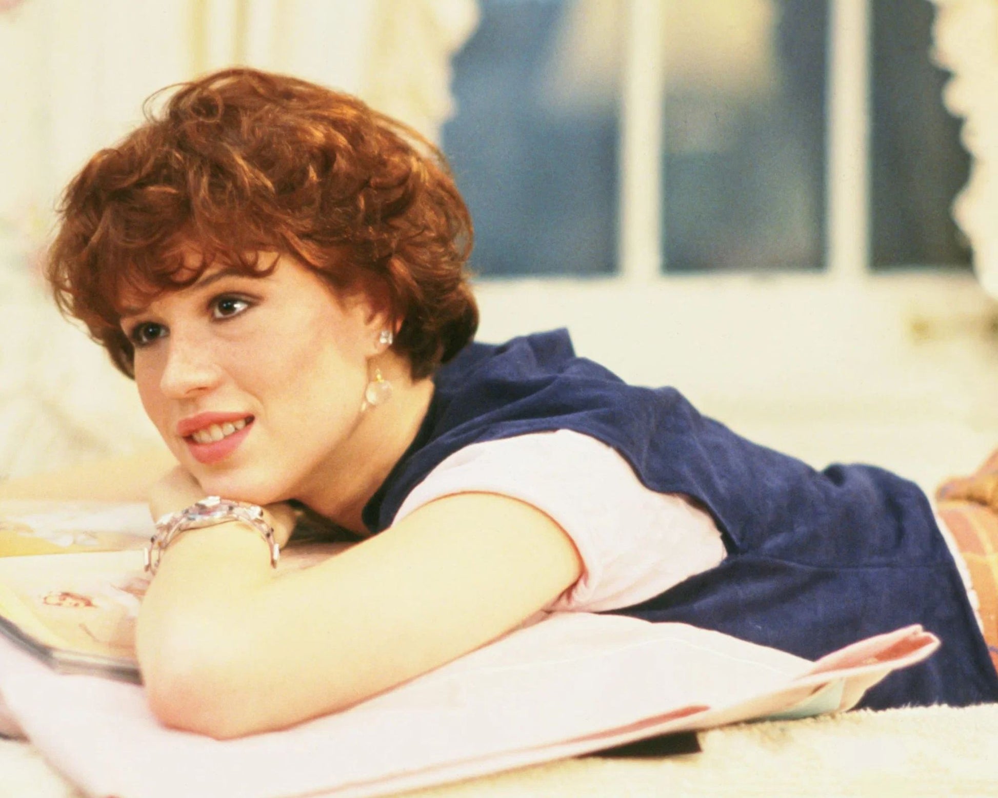 A woman with short curly red hair reclines on a bed or sofa, resting her head on her arms over an open book. She is wearing a navy blue top over a light pink shirt and has a silver bracelet on her wrist.